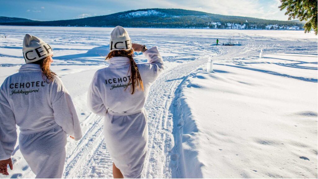 to kvinder i hvide badekåber og huer går i sneen ved Icehotel i svensk lapland