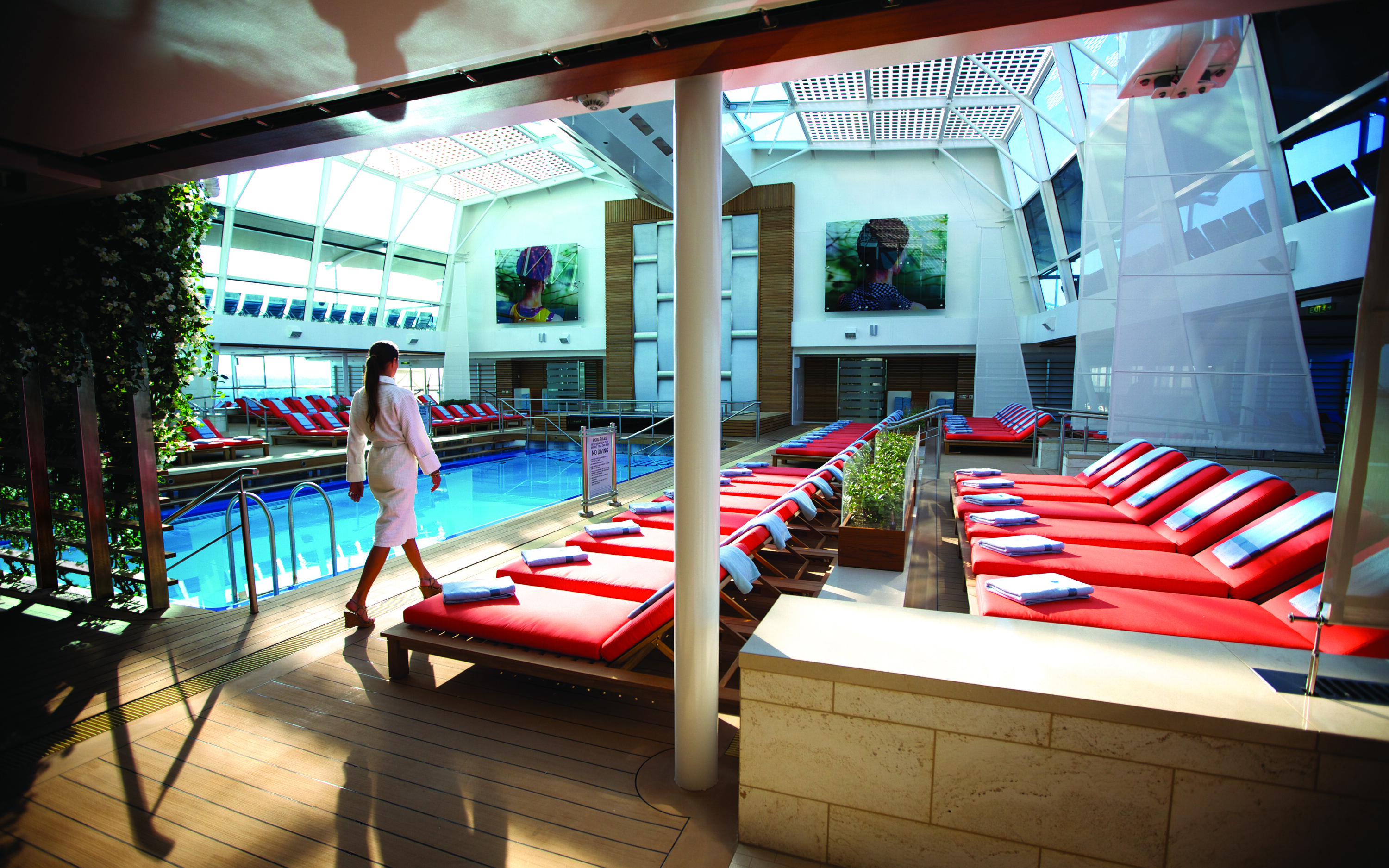Det indendørs Solarium på Celebrity skibet med azurblå pool, røde og hvide stribede liggestole og glasloft for optimal naturligt lys