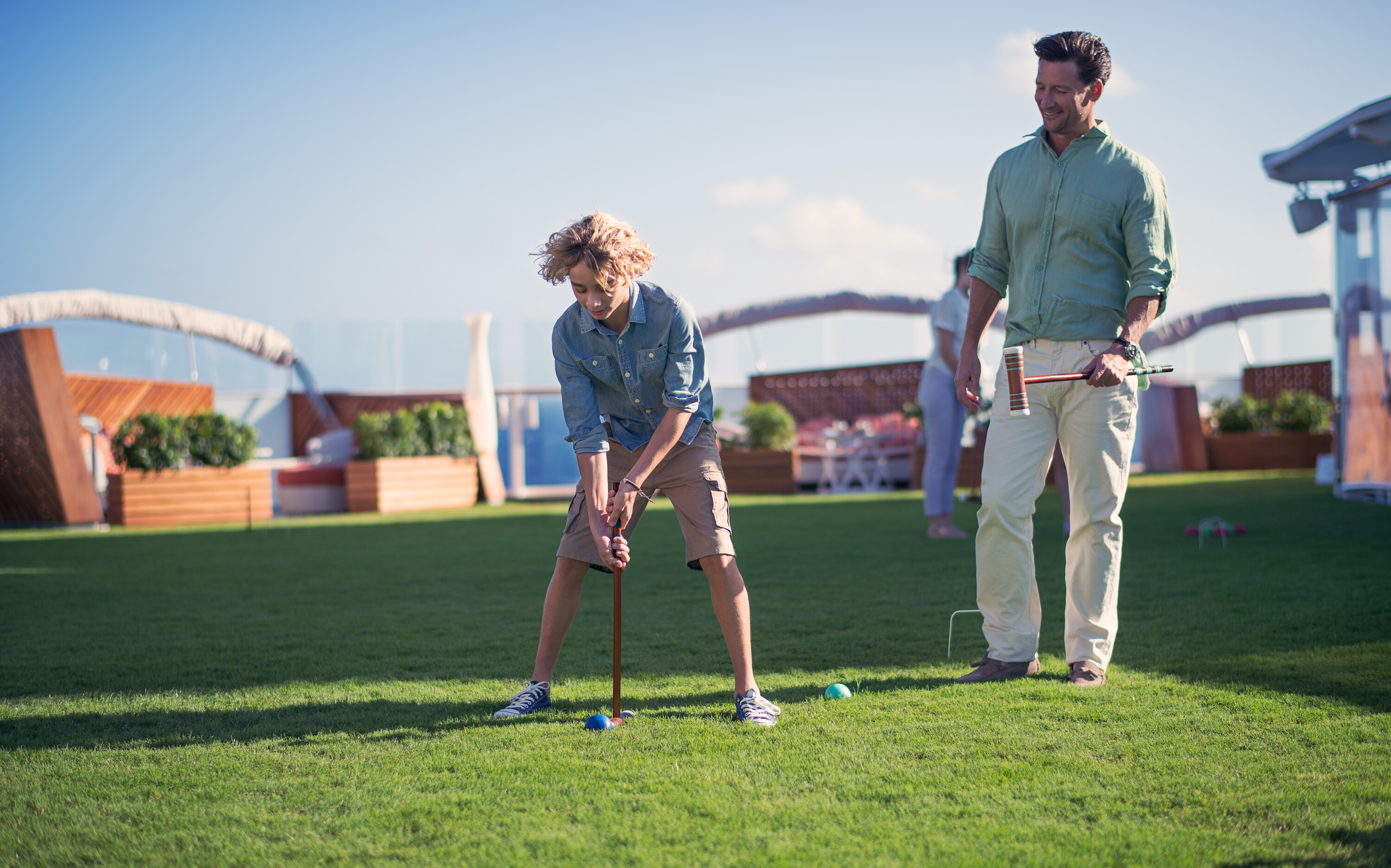 Far og søn spiller kroket på Celebrity skibets ægte græs Lawn Club under åben himmel med havudsigt og cruise faciliteter i baggrunden