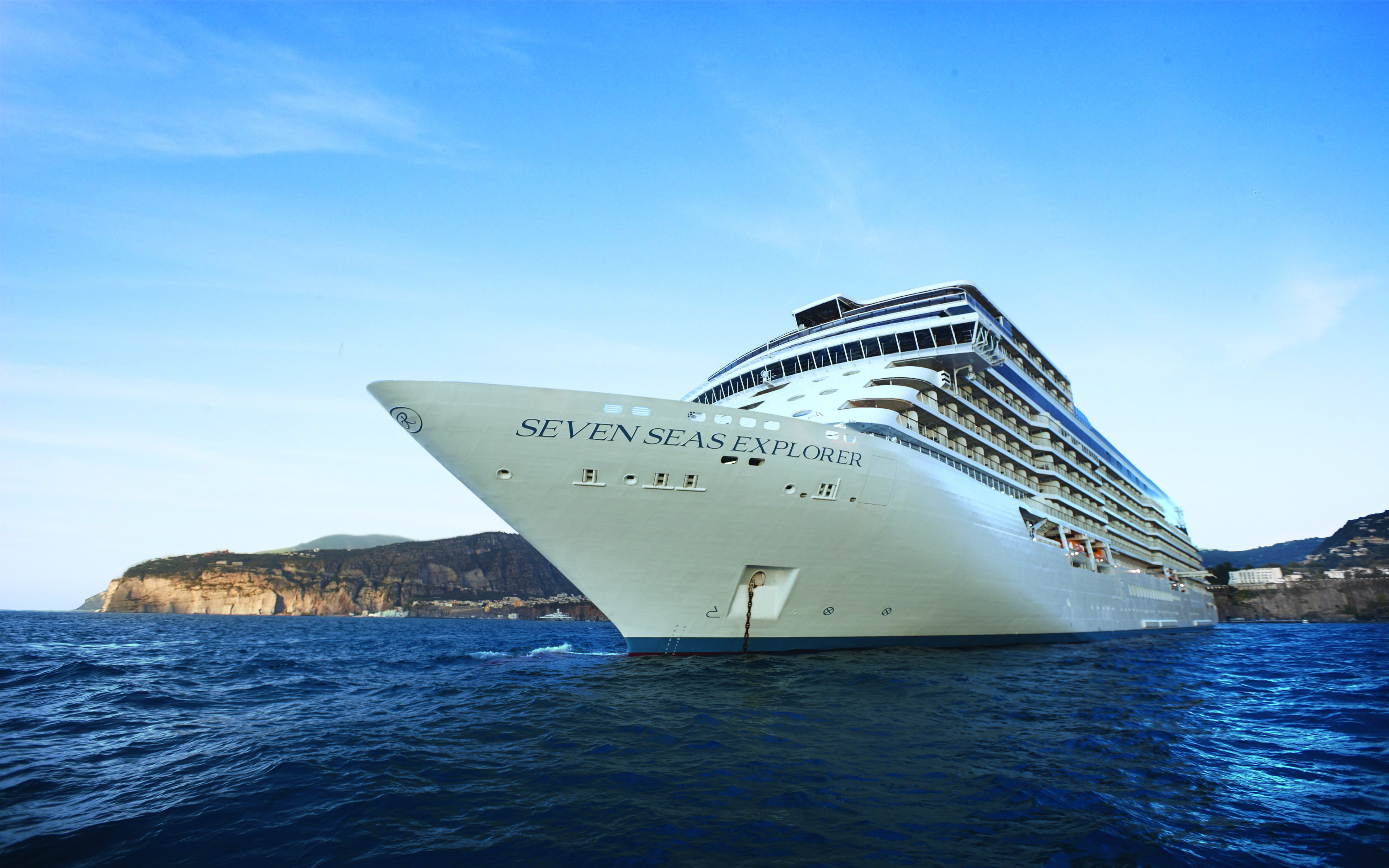 Krydstogtskibet Seven Seas Explorer set fra stævnen med kyst og klipper i baggrunden
