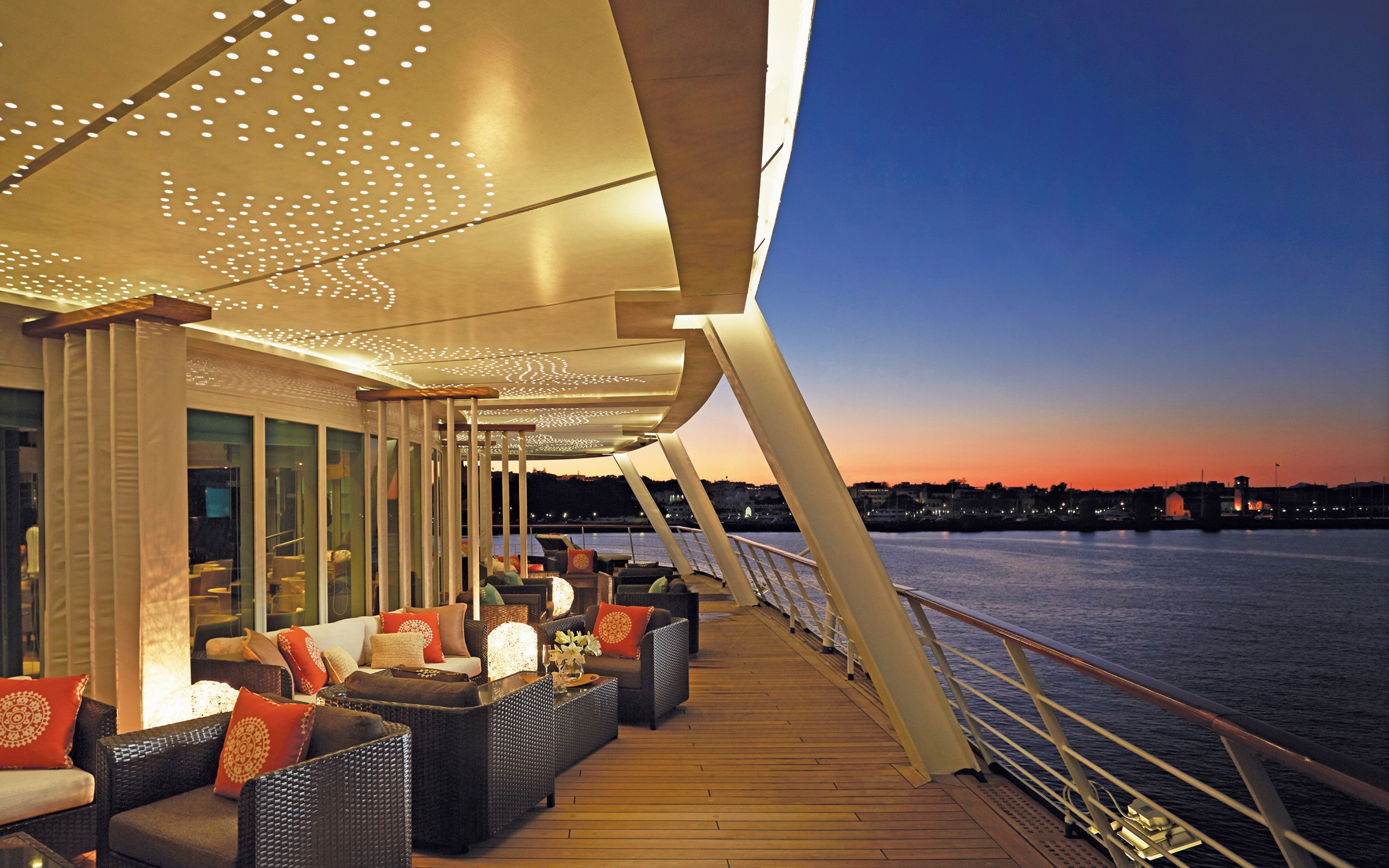 Stemningsfuld lounge på dækket af Seven Seas Voyager ved solnedgang over havnen